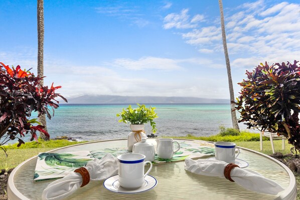 Uma mesa posta com xícaras e pires brancos, um vaso branco com folhas verdes e um porta-guardanapos branco com uma faixa marrom.