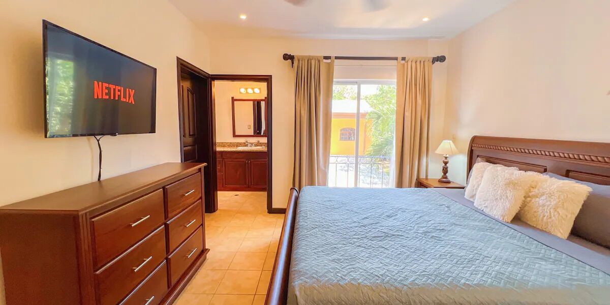 A hotel room with a bed, a TV mounted on the wall, a wooden dresser, and a bathroom visible through an open door.