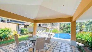 Uma área de varanda coberta com piscina, mobiliário externo e vegetação exuberante.
