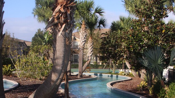 A clear, curved swimming pool with a tiled edge, surrounded by various trees and shrubs. - Upscale Kissimmee Retreat (Kissimmee)