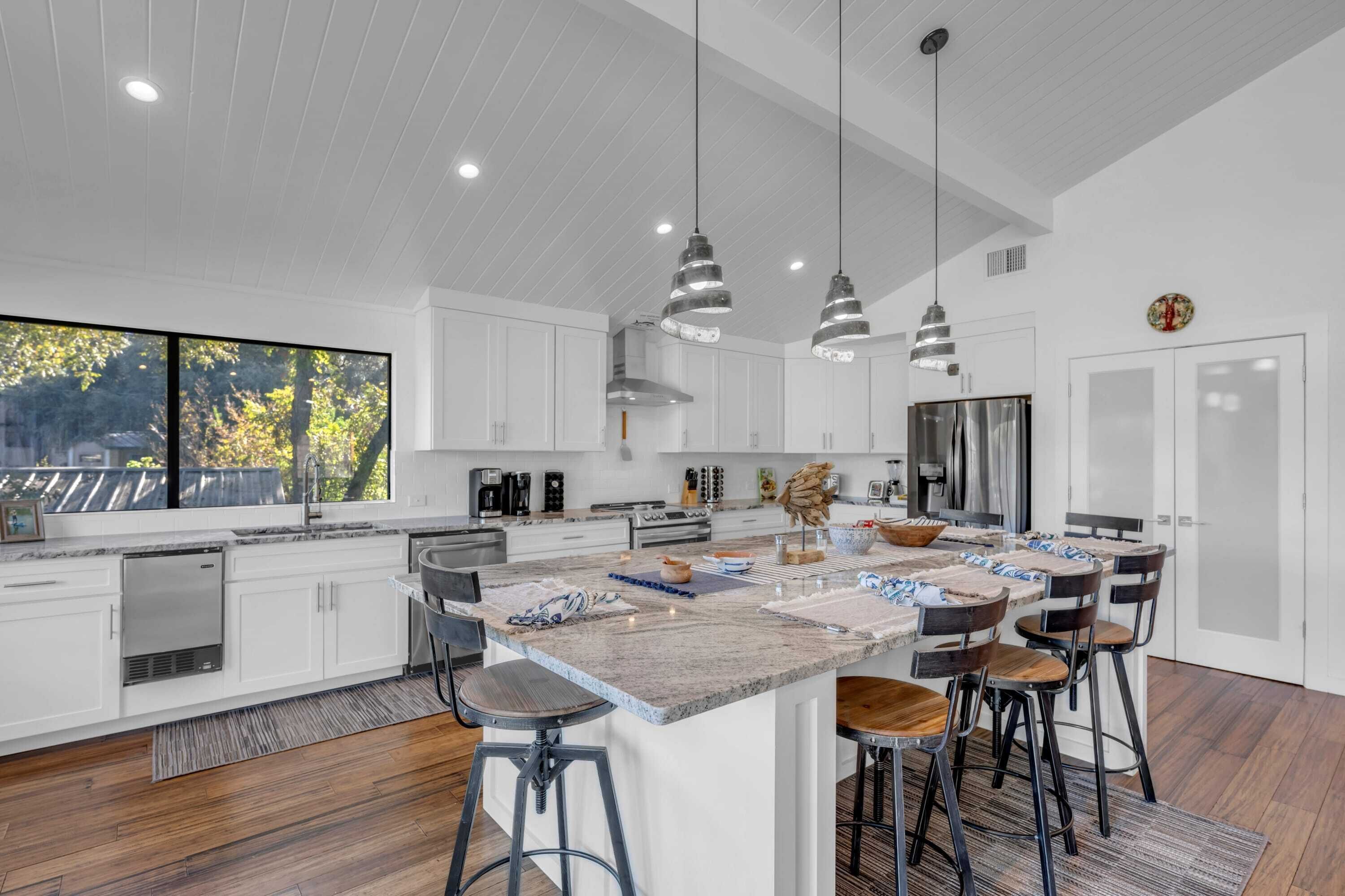 A modern kitchen with white cabinets, a large island with stools, pendant lighting, and a view of trees outside.
