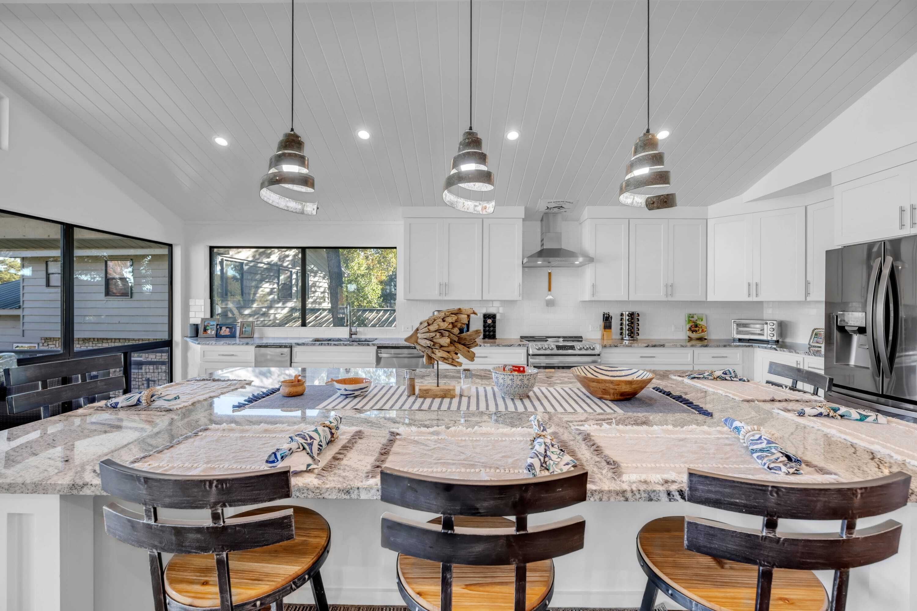 A modern kitchen with white cabinets, a central island with stools, pendant lighting, and a large window providing natural light.