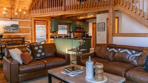 Een gezellig interieur van een chalet met een zithoek, twee leren banken, een salontafel met boeken en decoratieve objecten, een eethoek met een tafel en stoelen, en een keuken met groene kastjes en een houten trap.