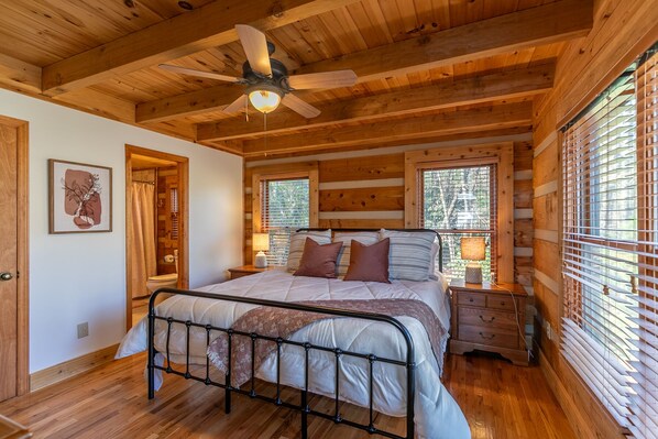 Une chambre dans une cabane en bois, avec un lit, deux tables de chevet, un ventilateur de plafond et une fenêtre avec des stores.