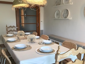 A dining area with a white table set for four, surrounded by wicker chairs. There is a woven light fixture above the table, and a shelf with decorative plates on the wall. - La Caseta de Quart, Girona (Quart)