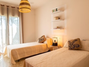 Een slaapkamer met twee bedden, een hanglamp, een plank met boeken en een mand, en een raam met gordijnen.