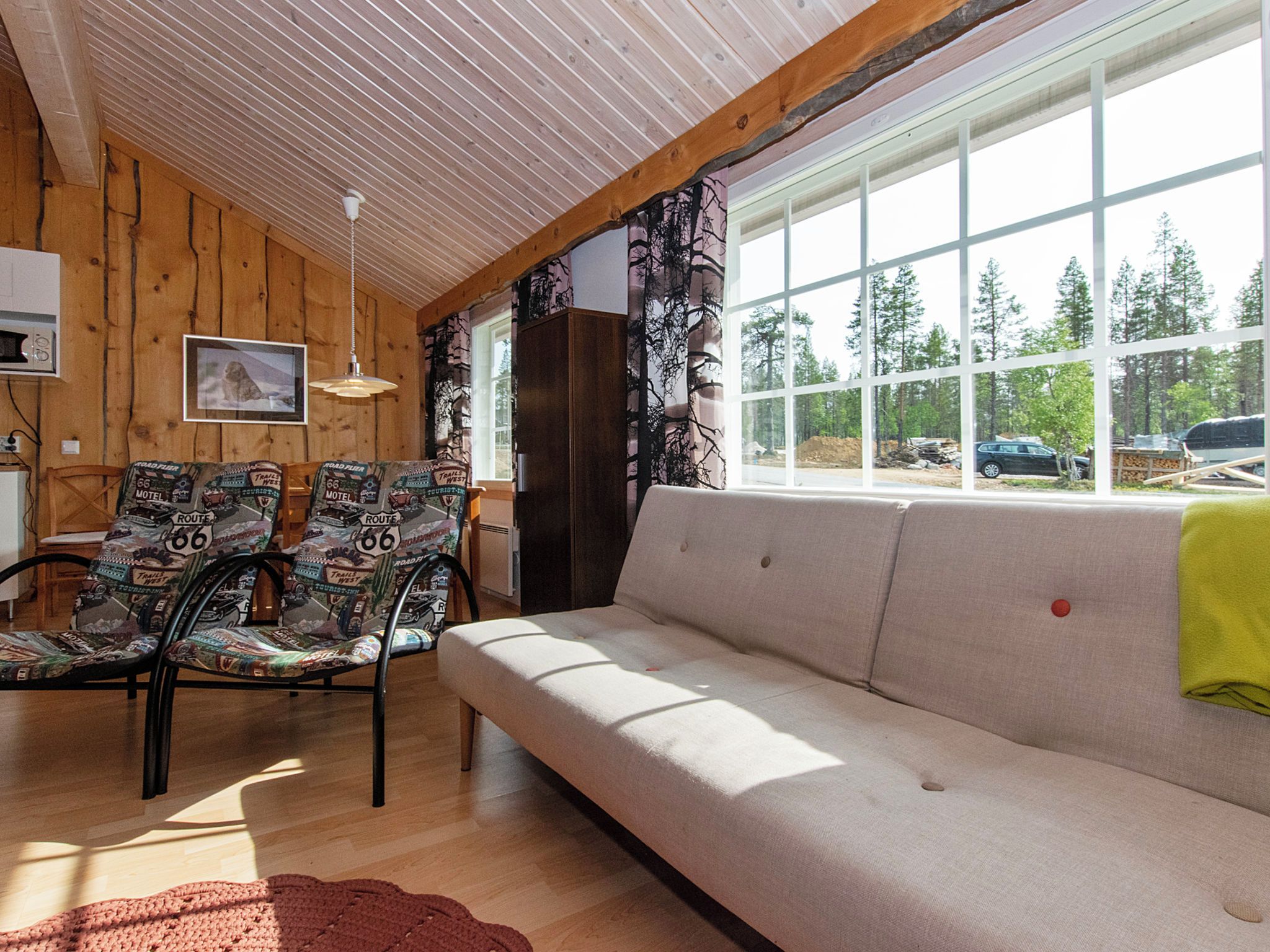 Una habitación con sofá, dos sillones, techo de madera, ventana con vista a árboles y un cuadro en la pared.