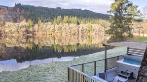 A lakeside deck with a hot tub, overlooking a calm lake and forested hills. - Woodhaven by Interhome (Invergarry)