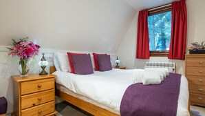 A bedroom with a bed, wooden nightstands, a dresser, a window with red curtains, and a vase of flowers. - Woodhaven by Interhome (Invergarry)