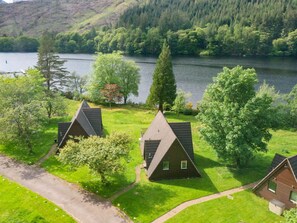 A lakeside property with three triangular-roofed cabins, a paved driveway, and a grassy area with trees. - Lothlorien Lodge 4 by Interhome (Invergarry)