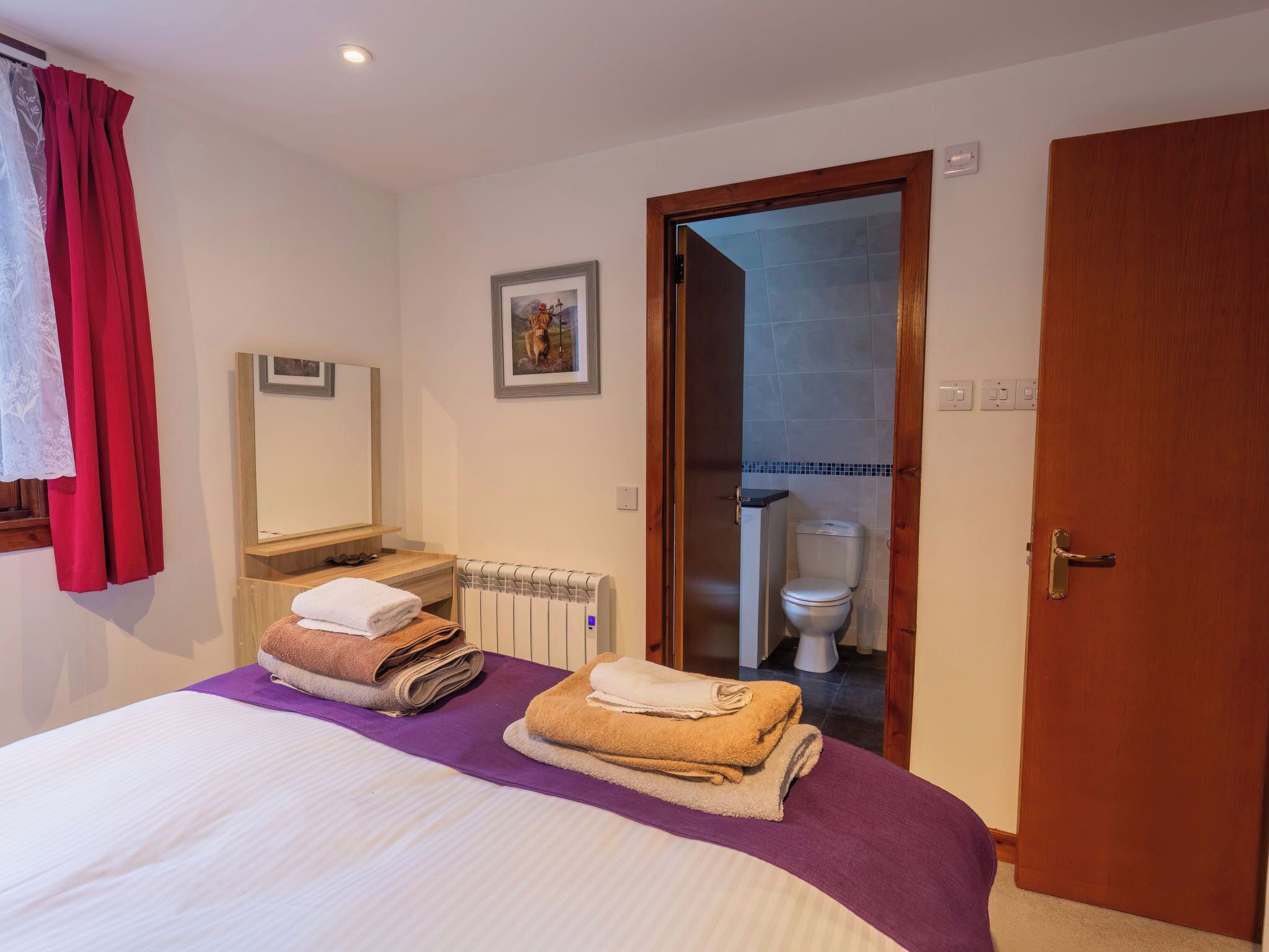 Habitación de hotel con cama, baño visible a través de una puerta abierta, un radiador y un cuadro en la pared.