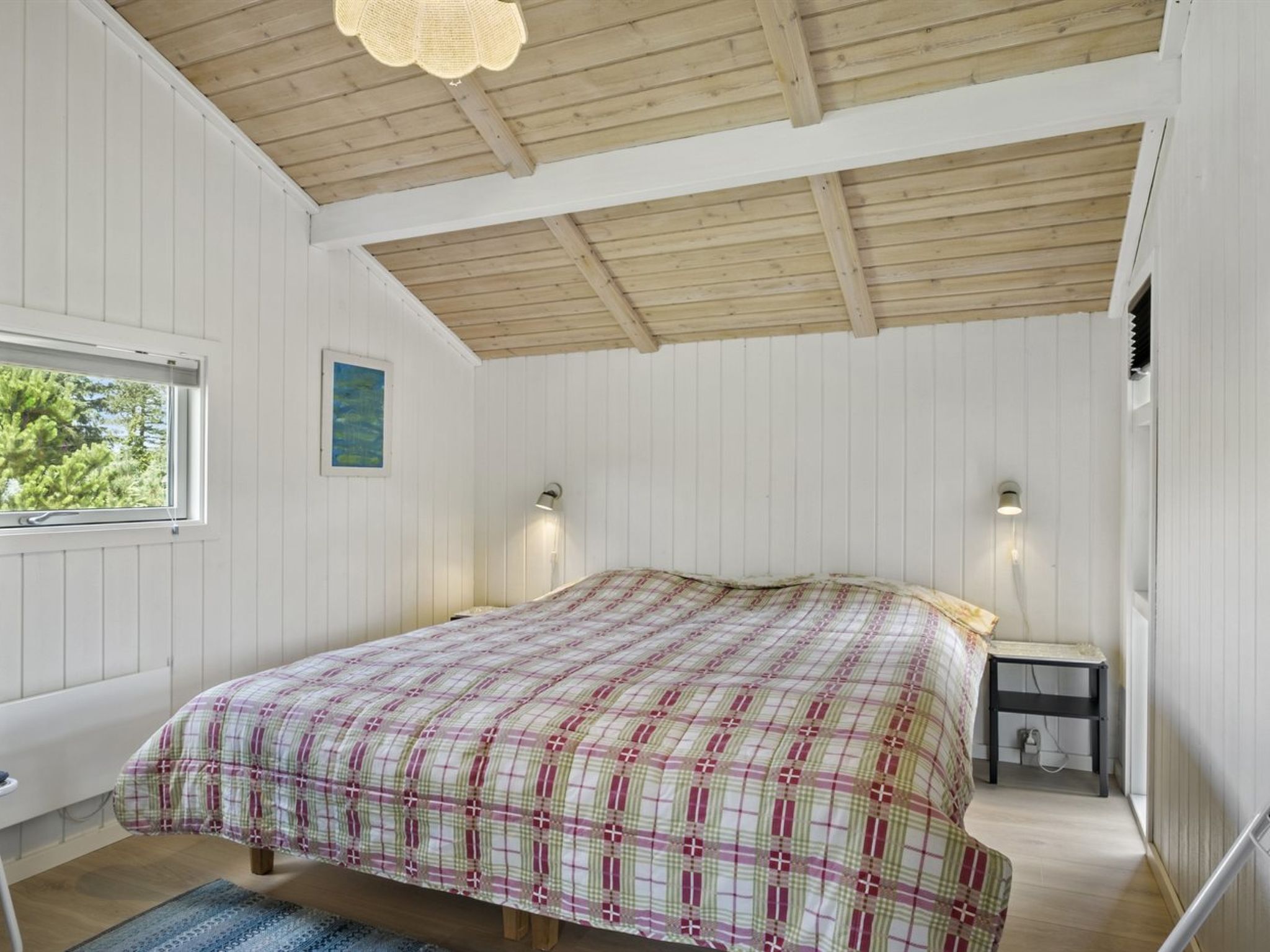 A bedroom with a wooden ceiling, a bed with a plaid bedspread, two bedside lamps, a window with a view of greenery, and a small table with a lamp.
