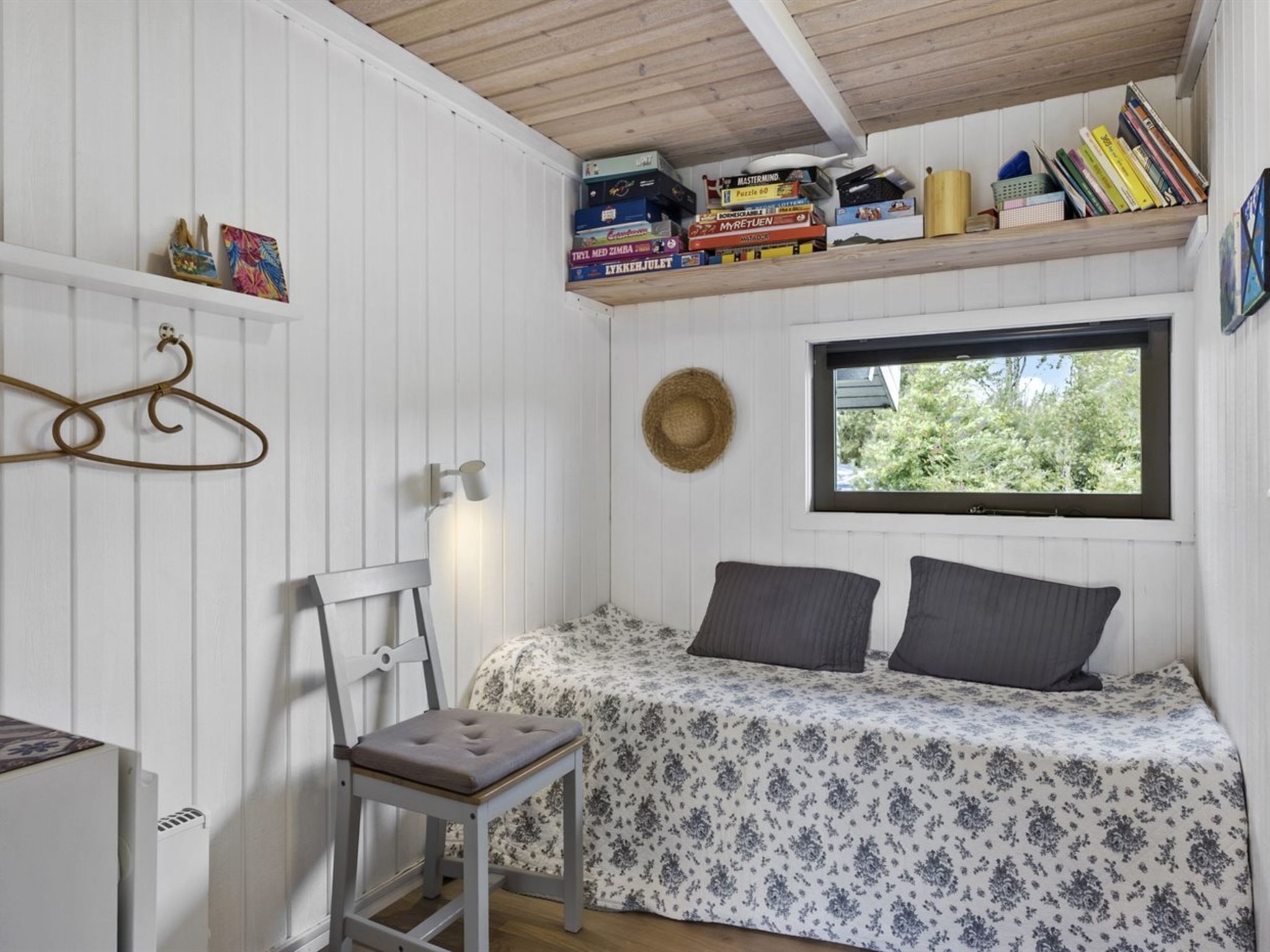 A small, cozy room with a bed, a chair, a hat on the wall, and a window with a view of trees.