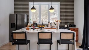 A modern kitchen with a central island, two pendant lights, a refrigerator, and a window with blinds.