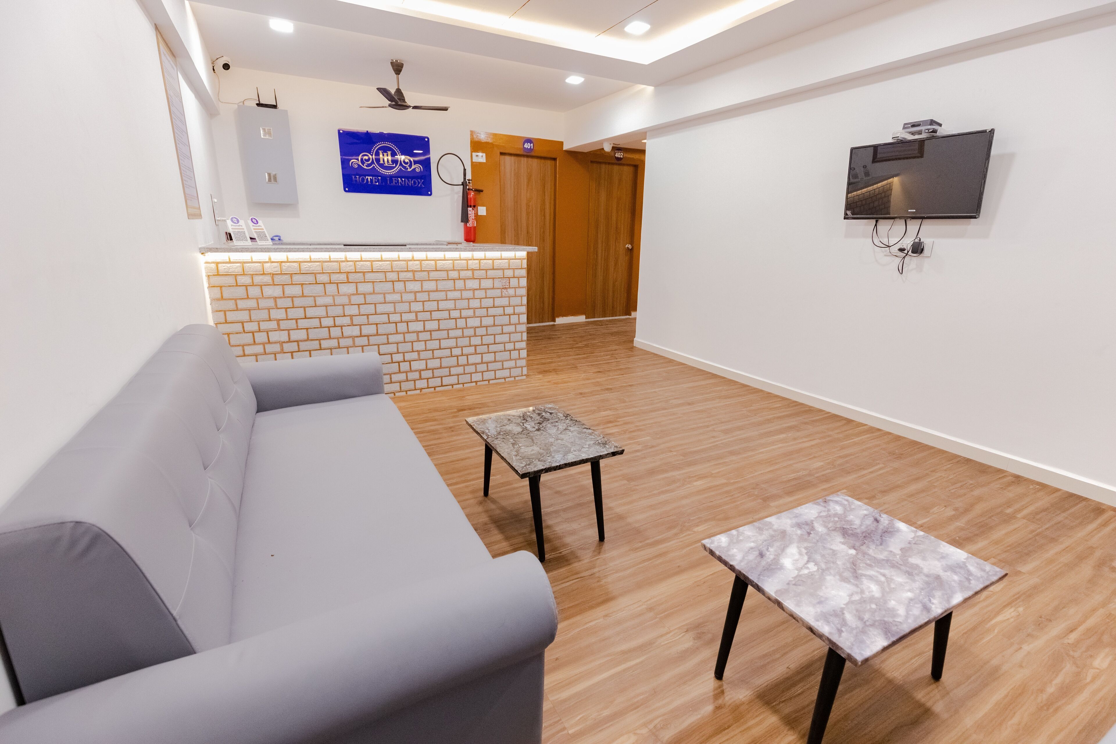 A modern living room with a grey sofa, two small tables, a TV mounted on the wall, and a reception desk with a blue sign.