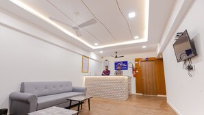A reception area with a grey sofa, a small table, a reception desk, a ceiling fan, and a TV mounted on the wall. - Hotel Lennox Best In surat (Surat)