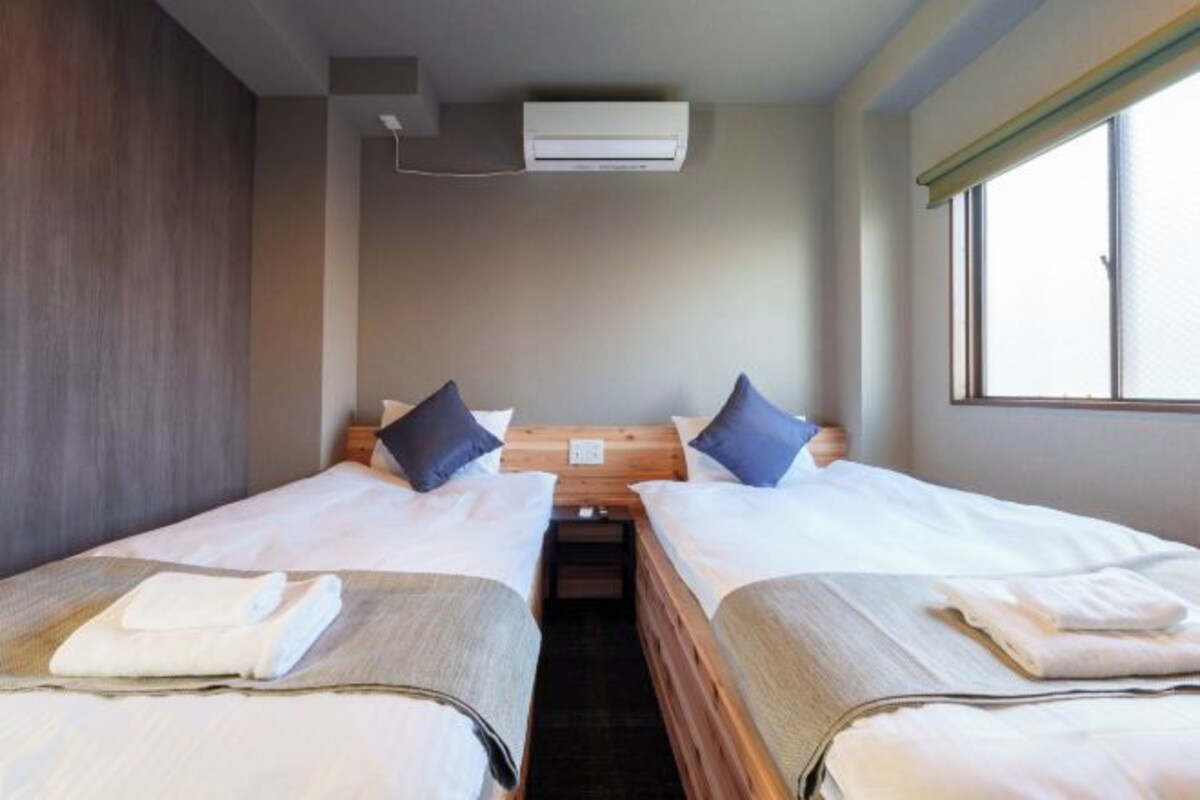 Ett hotellrum med två separata sängar, en sänggavel i trä, en luftkonditioneringsanläggning och ett fönster med persienner.