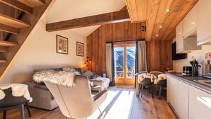 Una zona de estar acogedora con sofá y sillones, vigas de madera, una cocina con mesa de comedor y vistas a montañas a través de un ventanal.