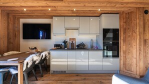 Cocina compacta con armarios blancos, horno integrado y televisor de pantalla plana montado en la pared.
