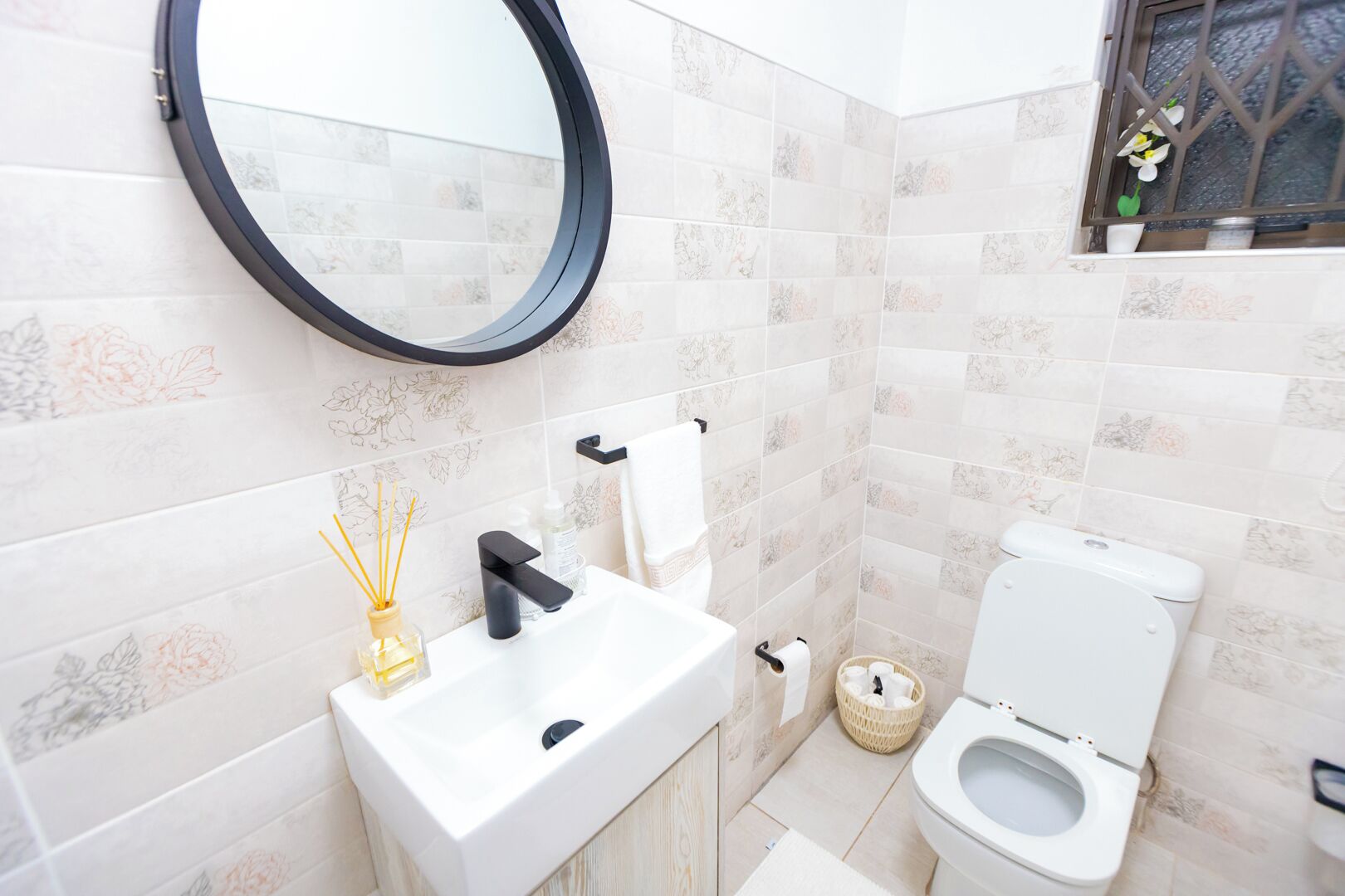 A bathroom with a white sink, black faucet, and a round mirror. There is a toilet with a white lid and a small basket beside it.