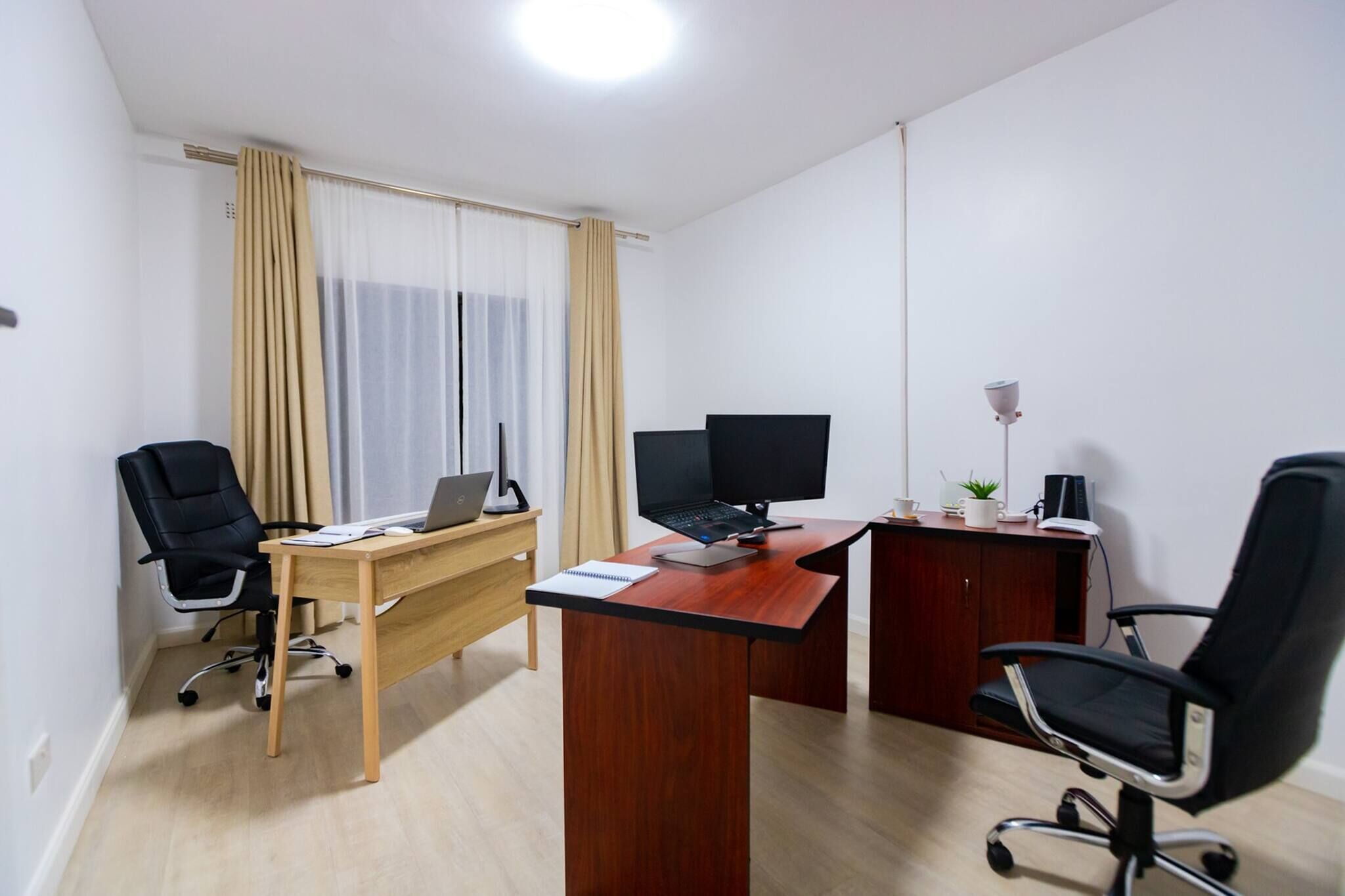 Een moderne kantoorruimte met twee bureaus, waarvan er één een laptop en de ander een beeldscherm heeft, een stoel en een klein tafeltje met een plant en een lamp.