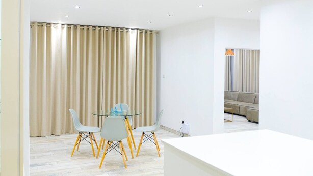Een moderne woonkamer met een bank, een glazen eettafel met stoelen en een groot raam met gordijnen.