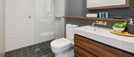 Un baño moderno con mampara de vidrio, inodoro blanco, tocador con espejo y una pequeña planta en maceta.