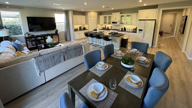 A modern living room with a sofa, a dining table set for six, and a kitchen in the background. - New 4B Home / Sequoia, EV, +More (Visalia)