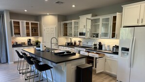 A modern kitchen with white cabinetry, a central island with bar stools, a built-in oven, and a refrigerator with a water dispenser. - New 4B Home / Sequoia, EV, +More (Visalia)