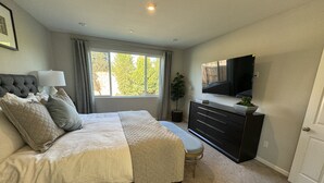 A bedroom with a bed, bedside table, dresser, TV, and a window with curtains. - New 4B Home / Sequoia, EV, +More (Visalia)