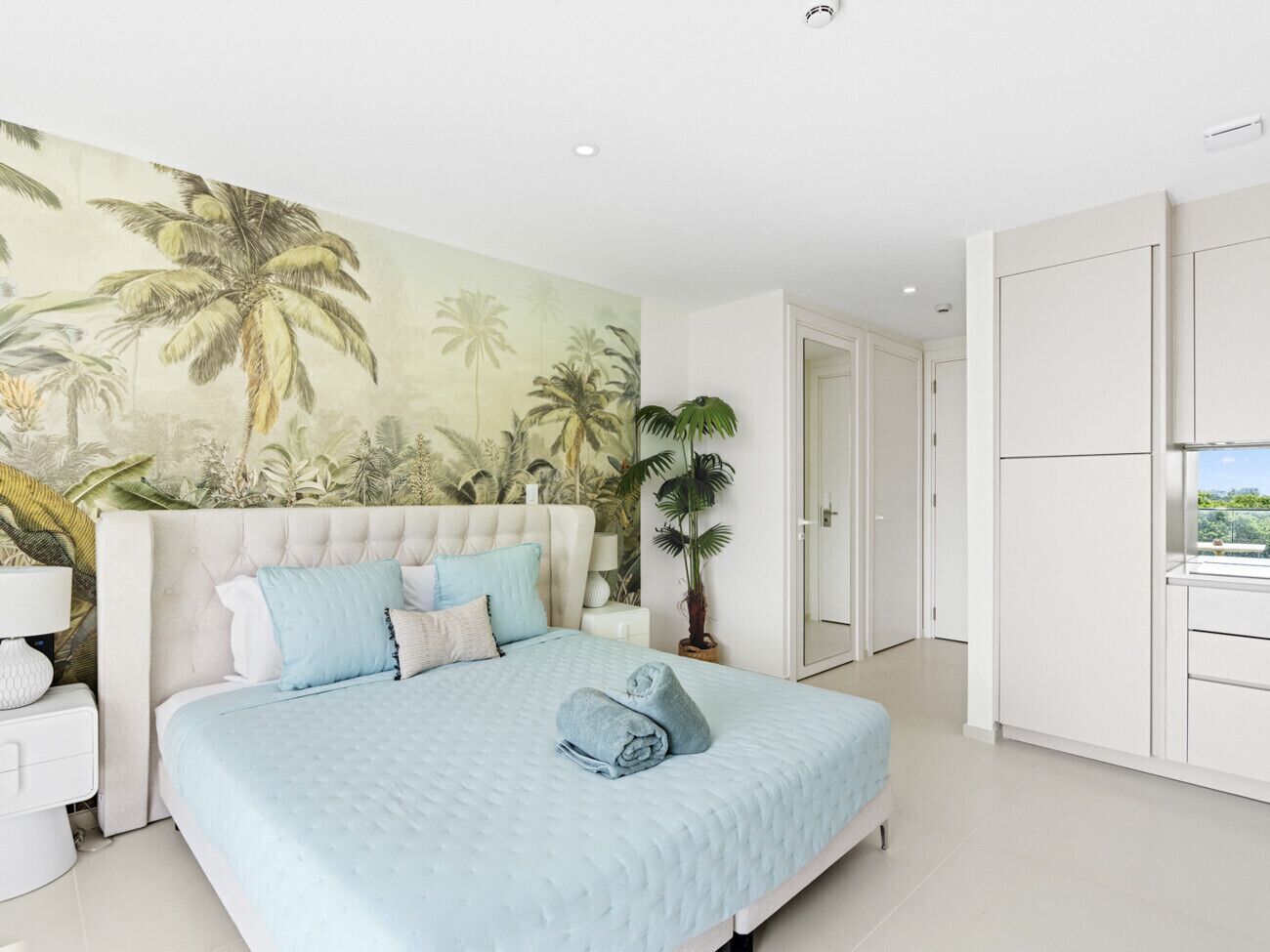 Une chambre à coucher comprenant un lit, une table de chevet, une fresque murale, une plante en pot et une armoire.