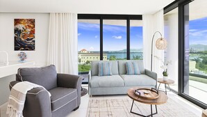 Una sala moderna con un sofá gris, un sillón gris y una mesa de centro redonda de madera. Se ve una ventana grande que ofrece vistas a un edificio y una masa de agua.