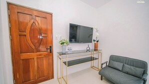 Une pièce avec une porte en bois, un téléviseur à écran plat fixé au mur, un canapé gris et une petite table sur laquelle est posé un vase de fleurs.