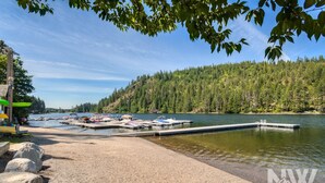 Ein Steg am See mit Booten, ein Sandstrand und ein bewaldeter Hügel.