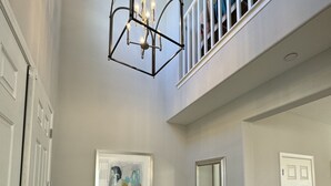 Un vestíbulo amplio con techo alto, una escalera a un nivel superior, una consola de madera con objetos decorativos, un cuadro abstracto enmarco y un espejo grande.