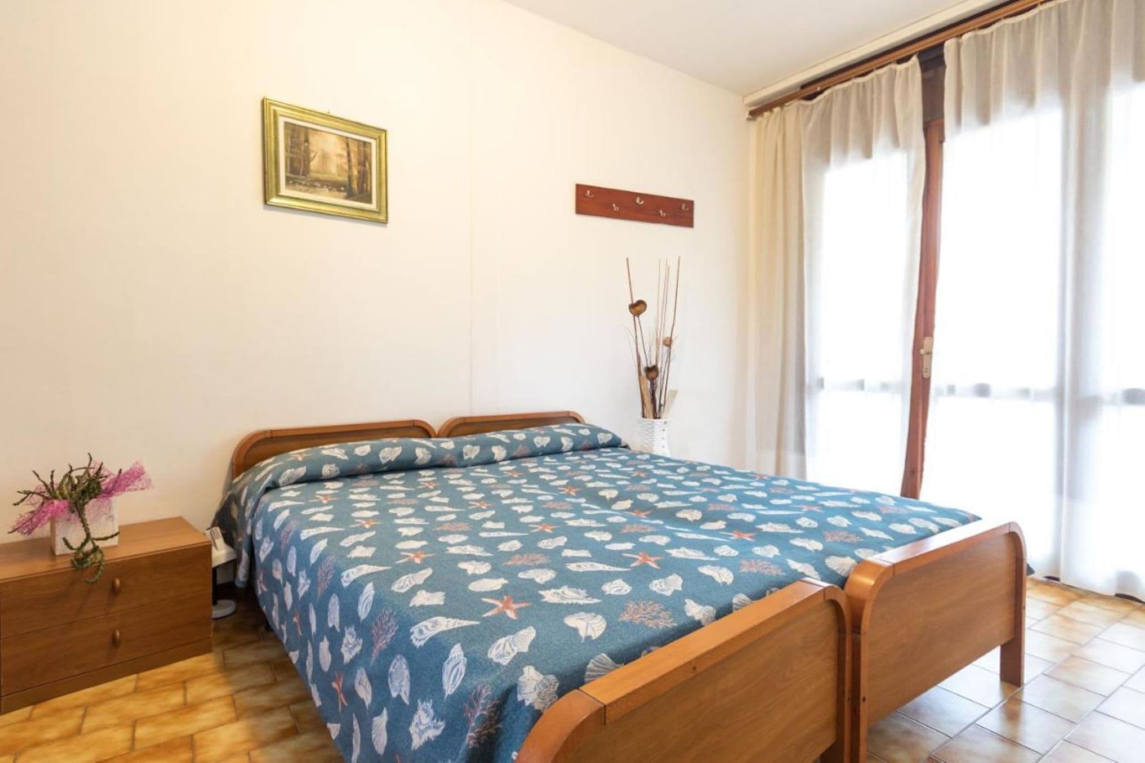 A bedroom with a bed, a nightstand, a wall-mounted shelf, and a window with curtains.