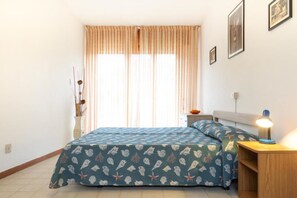 A bedroom with a bed, a bedside table with a lamp, a nightstand, and a window with curtains.