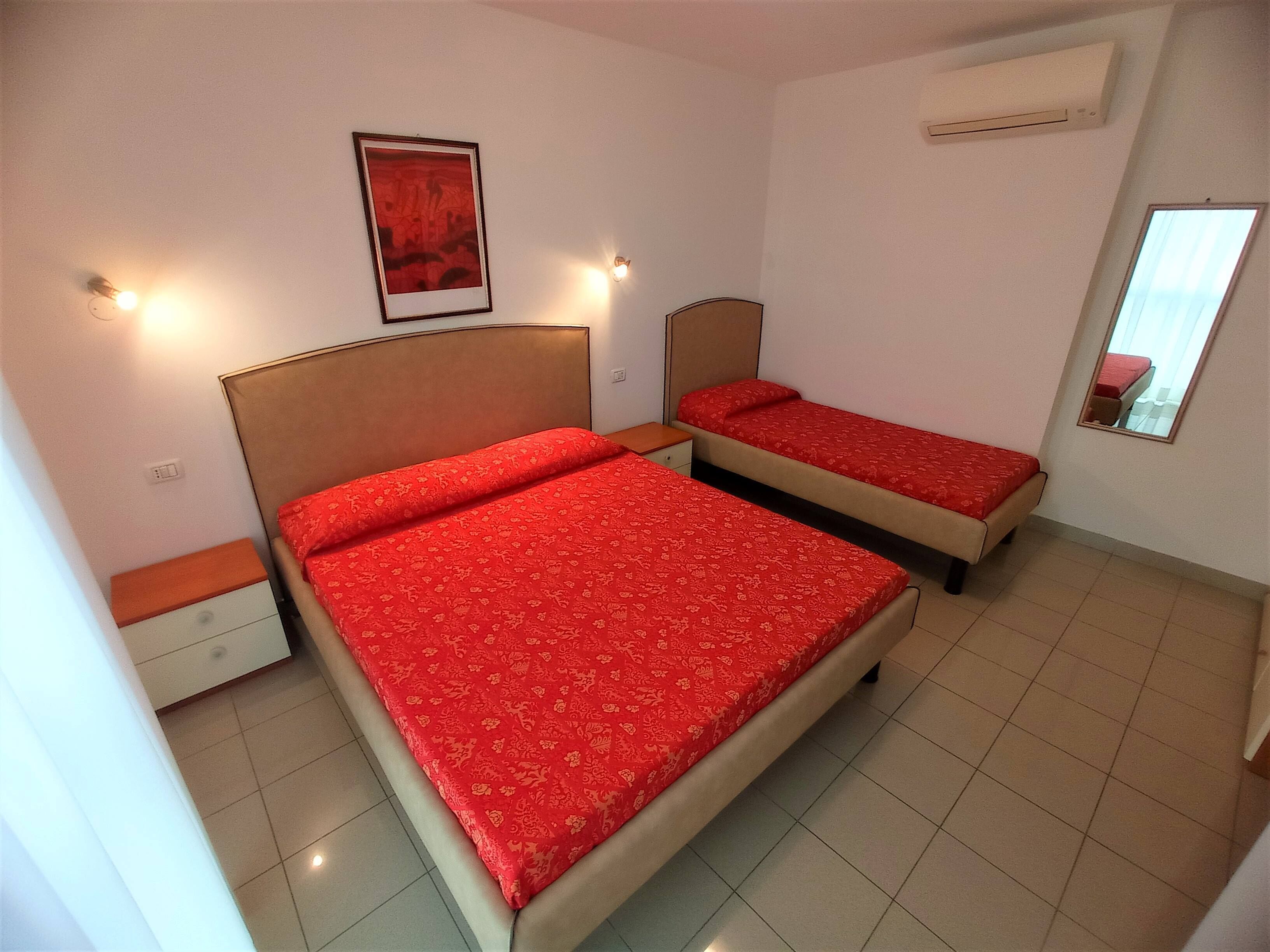 Habitación con dos camas, cada una con colcha roja, una mesita de noche, aire acondicionado de pared y un cuadro enmarcado.
