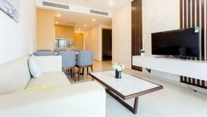 A modern living room with a sofa, dining table, chairs, a coffee table, a TV, and a kitchen in the background. - Garden View 2 bedrooms Ocean Suites (Da Nang)