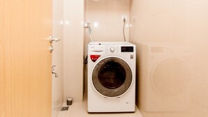 A compact, modern laundry room with a front-loading washing machine, a wooden shelf above, and a tiled wall. - Garden View 2 bedrooms Ocean Suites (Da Nang)