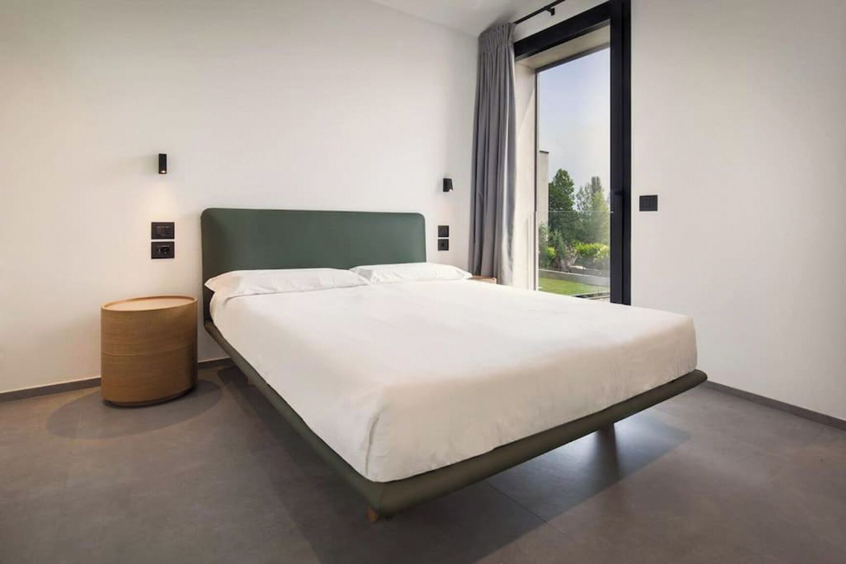 Quarto moderno com cama, luminária de cabeceira, um banquinho de madeira e uma grande janela com vista para o verde.