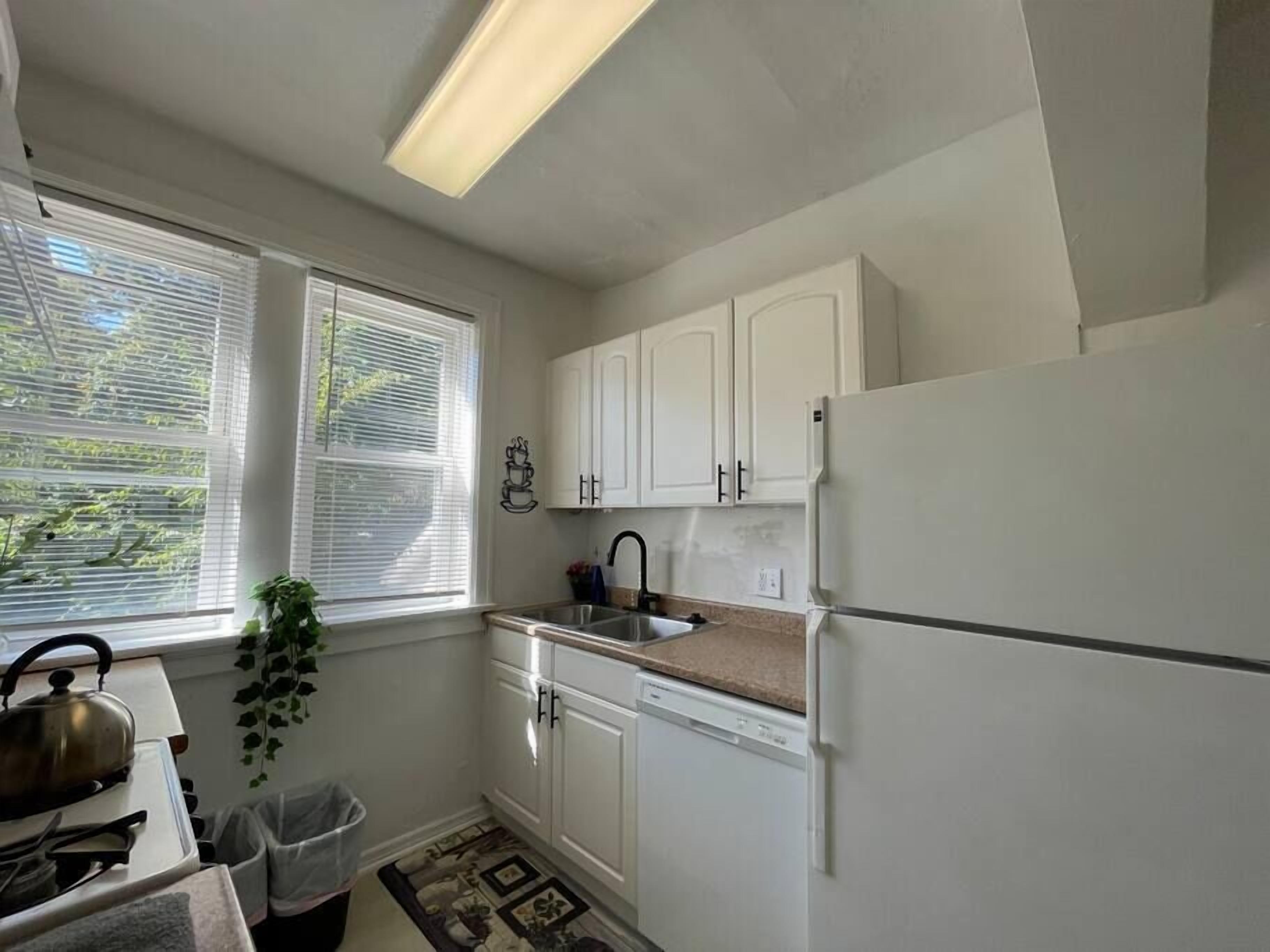 Eine Küche mit weißen Schränken, einem Spülbecken und einem Kühlschrank. Ein Fenster mit Jalousien und ein Topf mit Pflanze stehen auf dem Fensterbrett.