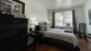 A bedroom with a bed, dresser, lamp, and a window with curtains. - 6653 1N | European Style Unit Near Forest Park (St. Louis)