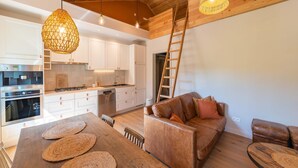 Una cocina moderna con armarios blancos, una mesa de comedor de madera con individuales tejidos y un sofá de cuero marrón con cojines.