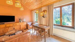 Una sala de estar acogedora con techo de madera, televisor de pantalla plana, mesa de comedor con sillas y un ventanal con cortinas.