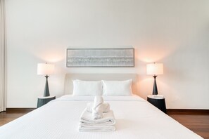 A neatly made bed with white linens and a folded towel in the center, flanked by two bedside tables with lamps, a framed artwork above the bed, and a plain white wall.
