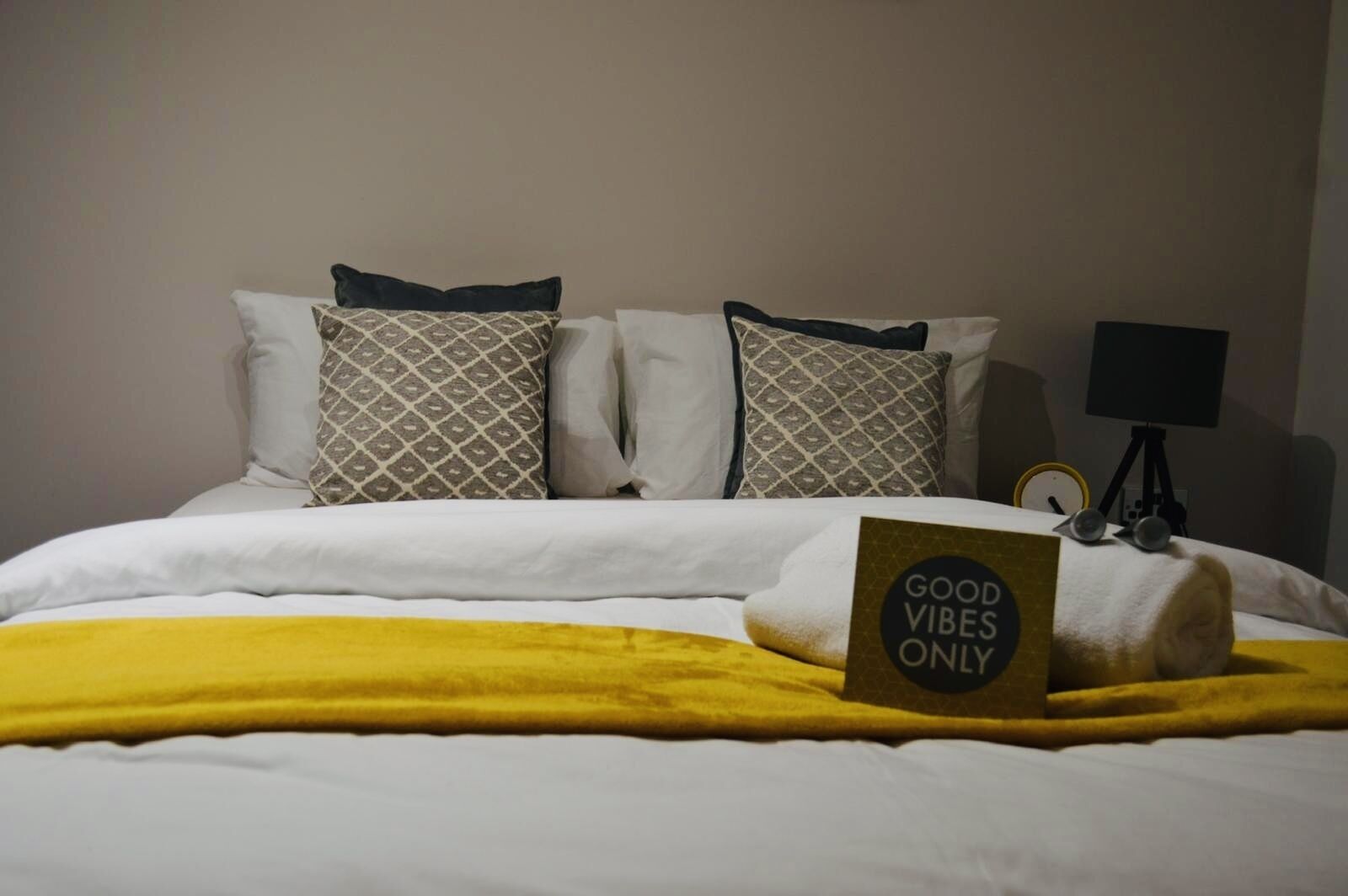 Un lit bien fait, avec une couverture jaune, des coussins à motifs et un panneau sur lequel on peut lire : « SEULEMENT LES BONNES VIBES ».