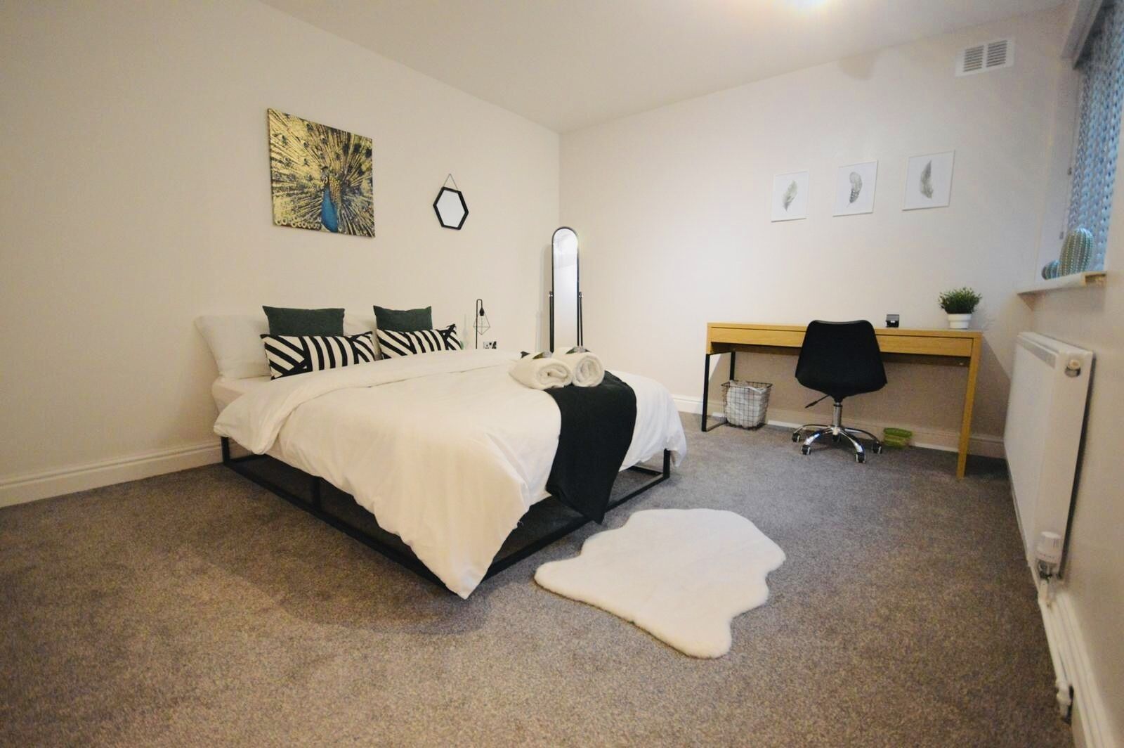 A bedroom with a bed, a desk, a chair, a mirror, and a peacock painting on the wall.