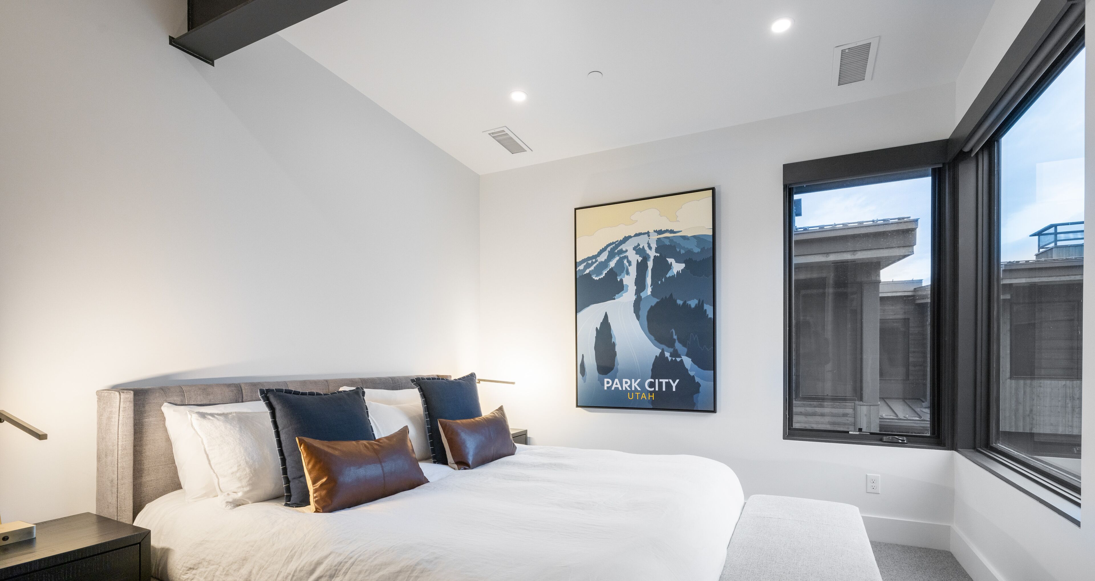 A modern hotel room with a large bed, a nightstand, a wall-mounted poster of a mountain, and a large window offering a view of the city.