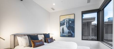 A modern hotel room with a large bed, a nightstand, a wall-mounted poster of a mountain, and a large window offering a view of the city.